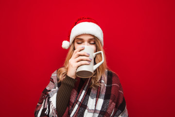 Cute girl in Santa hat and plaid drinks hot drink from big cup with closed eyes and enjoys on red background. Lady drinks tea and warms up in plaid in winter. Christmas concept.