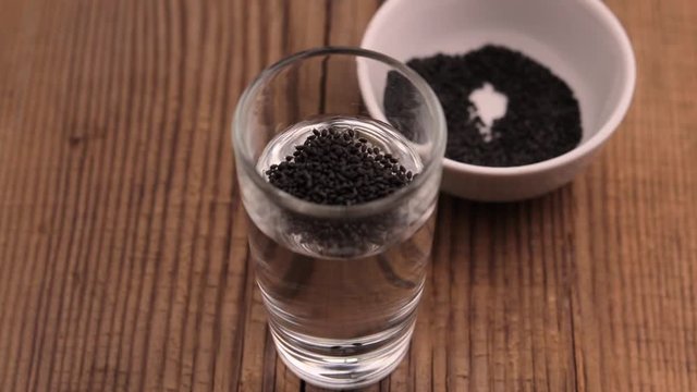 raw basil seeds are added with tea spoon to glass of water and mix on old rustic wooden table. Footage about superfood for cold beverages, smoothie and cocktails. Copy space.