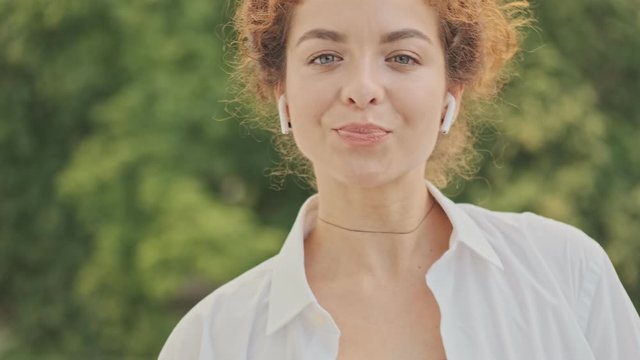 Attractive pleased young redhead woman in white shirt smiling and listening music with earphones while relaxing in the park