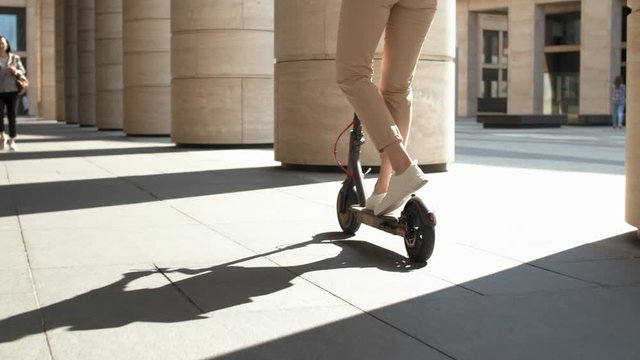 Rear view of young red-headed Caucasian woman wearing casual clothes, headphones and eyeglasses riding electric kick scooter in city and looking around
