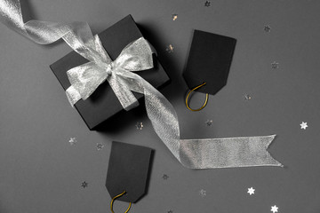 Gift box wrapped in black paper with silver ribbon, stars confetti and price tags on bright gray background. Copy space and top view. Black friday box gift present isolated.