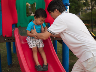 Asian child boy playing slider in playground together with dad in happy family time. Father support...