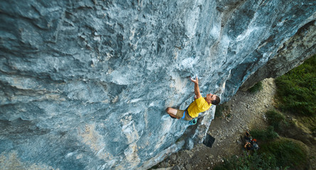 top view of man rock climber in yellow t-shirt, climbing on a cliff, searching, reaching and gripping hold. Conquering, overcoming and active lifestyle concept. - obrazy, fototapety, plakaty