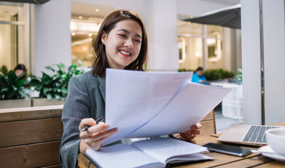 Cheerful female reading papers and working on laptop