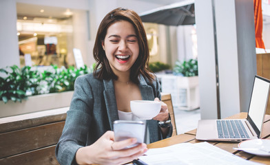 Young Asian laughing businesswoman with cup using smartphone