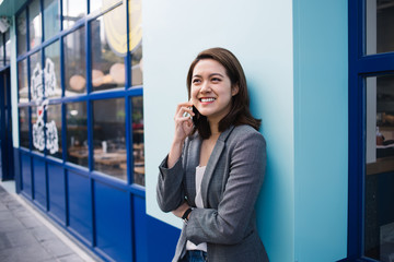 Young Asian smiling businesswoman talking on smartphone and leaning on blue wall