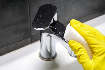 A hand in a yellow rubber glove wipes a dirty chrome metal faucet with a white melamine sponge....