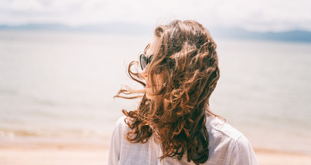 Young beautiful woman with dark curly fluttering hair with sunglasses on the seashore, closeup portrait