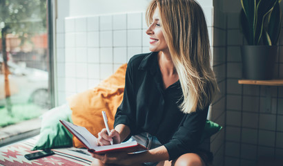 Cheerful Caucasian female blogger with blonde hair looking in window and smiling while creating...