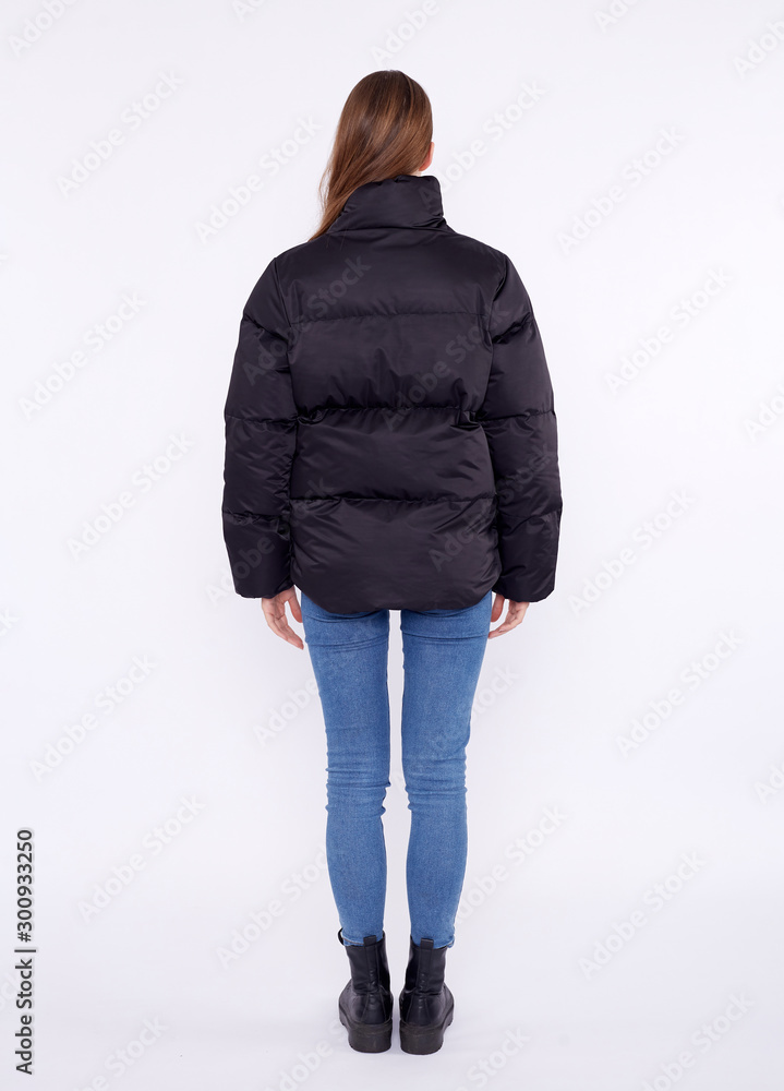 Wall mural Young girl in a black down jacket and blue jeans isolated on a white background.