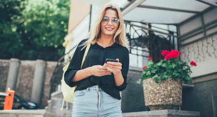 Half length portrait of cheerful hipster girl in trendy sunglasses standing outdoors and posing with cute smile on face,happy female traveller in casual apparel using modern cellphone for tracking gps