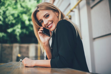 Portrait of happy female looking at camera while sitting at street cafeteria and using public internet for making international call, cheerful woman contacting friends via roaming wireless and app