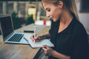 Side view of positive skilled female student making notes of information from useful website in...