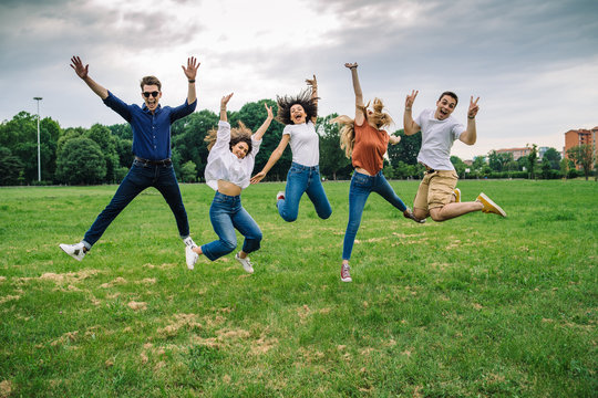 Group of five friends jump into the air with their hands up - Millennials have fun together at the park in the summer