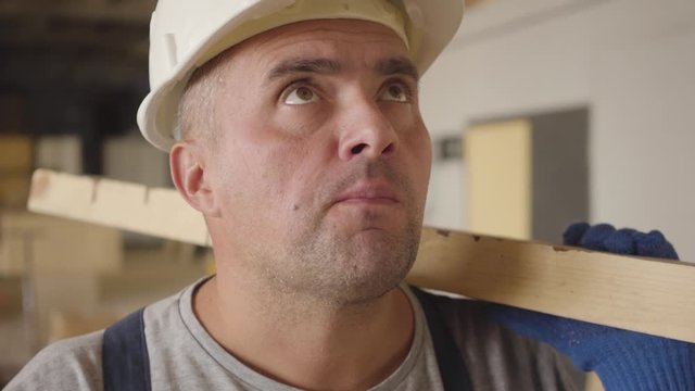 Close-up face of a Caucasian builder with brown eyes eating bun at the construction site. Strong muscular worker having lunch at work. Man in white helmet holding wooden plank and chewing.