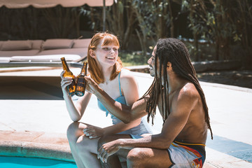 Multiracial couple sitting on the edge of the pool toasting with beer and smiling with the garden in the background