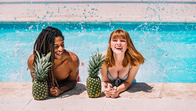 Multiracial couple leaning on the edge of the pool splashing with their feet and with two pineapples