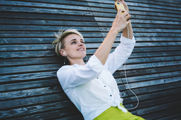 Happy woman dressed in stylish apparel enjoying leisure time for listening favourite positive music via electronic earphones and mobile application while clicking selfie photo on front camera