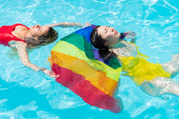 Two girls from different ethnic groups relaxed floating on the surface of the water sunbathing with an lgtb flag between the two