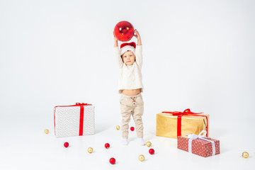 Little boy standing between gifts and holding big red Christmas ball in hands. Isolated on white background. Holidays, christmas, new year, x-mas concept.