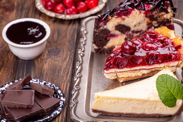 Chocolate cherry fruit cake, cherry cheeseccake and cheesecake on silver tray. Three pieces of sweet pies on wooden  white background
