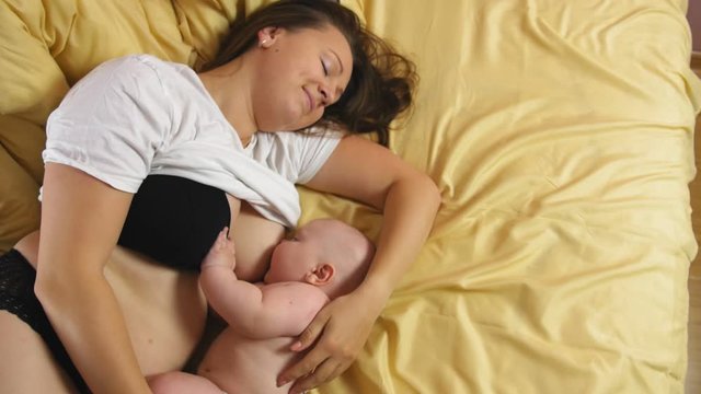 Young mother breastfeeds her baby while lying on a sofa. The camera shoots from above. Medium shot.