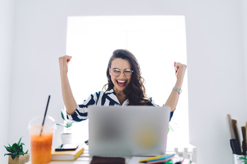 Joyful young woman sitting at table with hands up