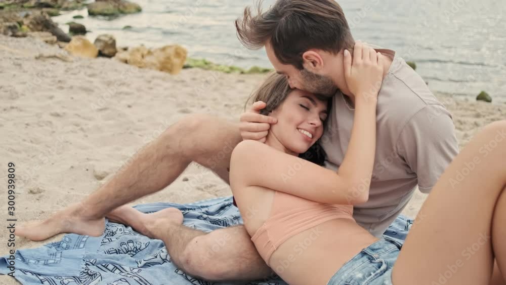 Wall mural Handsome calm young bearded man hugging with his cute young girlfriend and kissing her in forehead while lying on plaid near the ocean