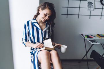 Cheerful brunette in stylish striped dress reading book and smiling