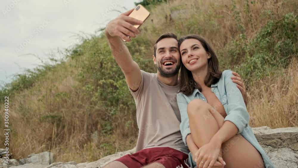 Poster Attractive cheerful young lovely couple hugging and taking selfie photo on smartphone while sitting outdoors on green hills