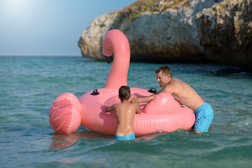 Active European boy nd his dad swimming in ocean with inflatable pink flamingo. They have real fun during their summer holidays. - 300941468