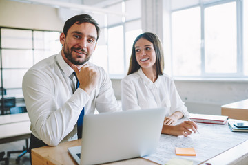 Portrait of successful positive male professional architect sitting at desktop with laptop computer and sketches and looking at camera while smiling female colleague near working on blueprints