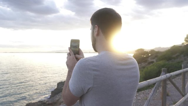 A man takes pictures of the sunset with the cell phone on the coast, the sea and the golden sky are seen while he remains behind a fence inside a rural road