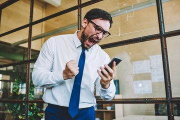 Young aggressive male trader emotionally reacting to online change in financial market and screaming during watching on telephone.Angry man shouting to smartphone while watching football match