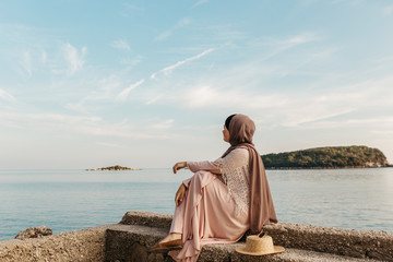 portrait of young European Muslim women with hijab sitting on the beach with hat in her hand. She is happy and relaxed. Sea is in the background. She is looking in the distance.