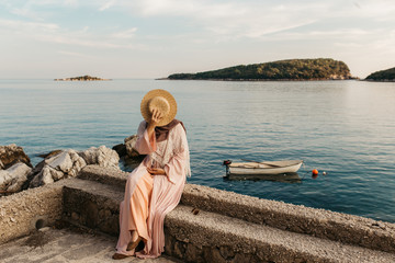 portrait of young European Muslim women with hijab sitting on the beach with hat in her hand. She is happy and relaxed. Sea is in the background. She is looking in the distance.