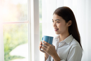 Asian woman drinking a cup of coffee and looking through the glass window after wake up in the morning