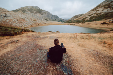 Stock aerial photo of a photographer girl from behind sitting in front of a lake surrounded by mountains with a camera in his hands. Travel and nature