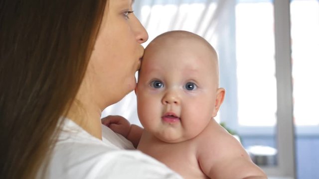 Mother holds a chubby smiling baby in her arms at home. The kid is looking at the camera. Close up shot.