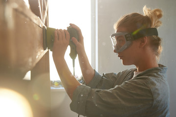 Young woman in safety glasses polishes a wooden bed with random orbit sander, DIY concept