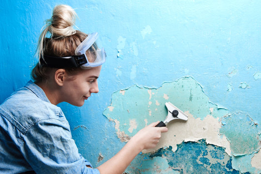 Girl With Scraper In Hand Peels Off A Layer Of Cracked Blue Paint On The Wall In The Apartment, Woman Makes Repairs