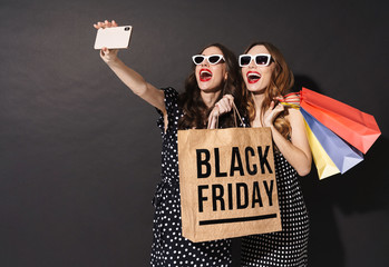 Image of girls taking selfie on cellphones and holding black friday bag