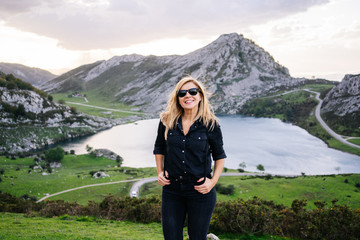 Naklejka na ściany i meble A beautiful caucasian blond woman in front of the camera wearing casual clothes in a mountainous landscape with lake