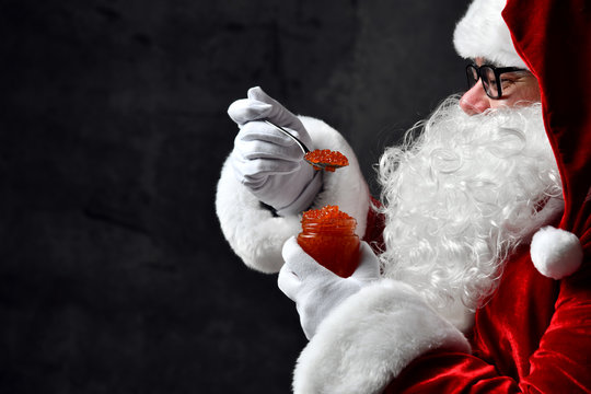 Santa Claus eating holding spoonful of red caviar fish salmon on dark gray concrete wall. New year and Merry Christmas
