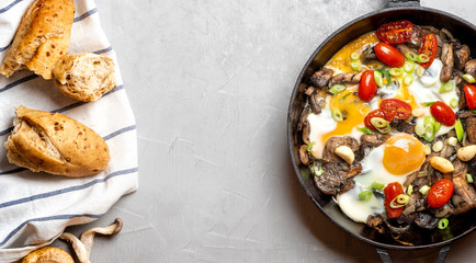 Tasty and hearty breakfast or snack. Fried eggs with mushrooms, tomatoes and onions in a pan on a grey background, top view, free space for text.