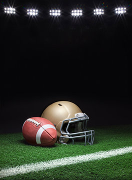 A gold football helmet and football on a grass field with stripe on dark background with stadium lights