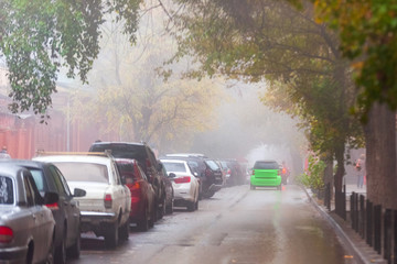 Morning fog in the city in the fall. A small green car is driving along a narrow street. There are a lot of parked cars along the road