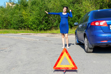 Photo of young woman with raised hand standing near broken car with open hood. Car insurance concept