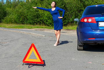 Photo of young woman with raised hand standing near broken car with open hood. Car insurance concept