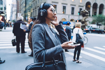 Attentive serious female in eyeglasses and headphones with mobile standing on street